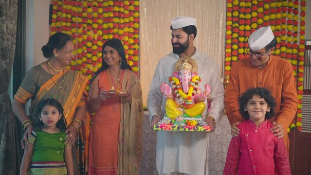 Happy ethnic Hindu Indian young man holding a Ganapati idol with smiling family members in traditional clothes during the Ganesha Chaturthi festival or celebration in a house decorated with garlands