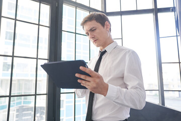 Fototapeta premium A businessman working on his digital tablet. A serious man at the screen in the office. The concept of a work routine. The background with the window with copy space. High quality photo
