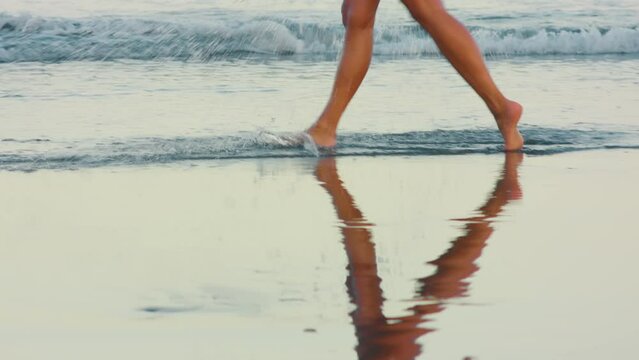 Women's feet walk on the sand near the sea with waves. The surf, a wave with foam washes the woman's feet. 
Female feet go along the shore near the water.