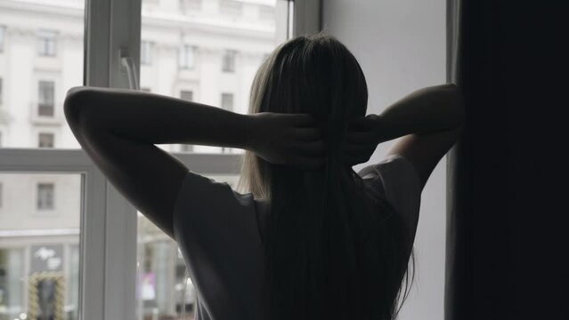 A young woman woke up, stands at the window, stretching her hands behind her head, looks out the window overlooking the city streets, the building. Close-up