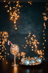 Seasonal greetings Christmas card. New Year's still life. Vintage wooden table, large wine glass filled with Christmas balls. Dinner served Santa Claus centerpieces and bokeh lights against dark wall.