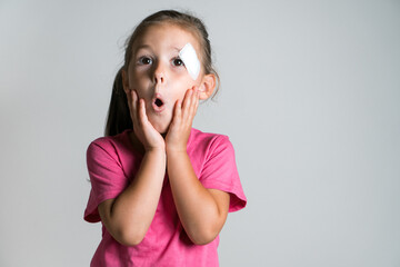 Close up surprised, shocked adorable dark haired child, girl with ponytail with scratch and patch on face, with arms on face, wear pink shirt on white background. Wounds, fidget children. Copy space