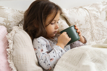 Sweet child, little dark haired preschooler girl drink hot unpalatable medicine from green mug, lying in pajamas in soft bed. Recovering, health care. Taking medicine, sore throat, cough and pain