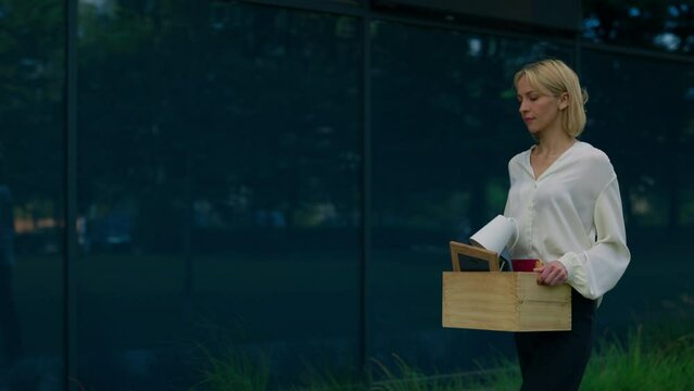 Side view of the jobless blonde woman who has just been released and going near the work place and sadly looking, holding a box with work items .Jobless people concept.