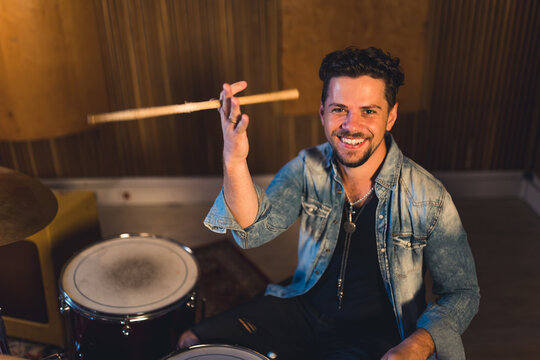 medium shot of a young drumer man holding drums sticks and smiling widely, musical band concept musical studio. High quality photo