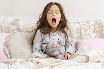 Funny, cute, tired little brunette girl yawning with open mouth, carrying teddy bear toy, sit on...