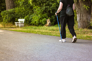 Nordic walking with sticks, a type of physical activity for retirees. An elderly middle-aged woman does Scandinavian walking outside in a summer park on nature. Sporty active faceless woman back view.