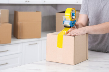 Man taping box with adhesive tape dispenser in kitchen, closeup