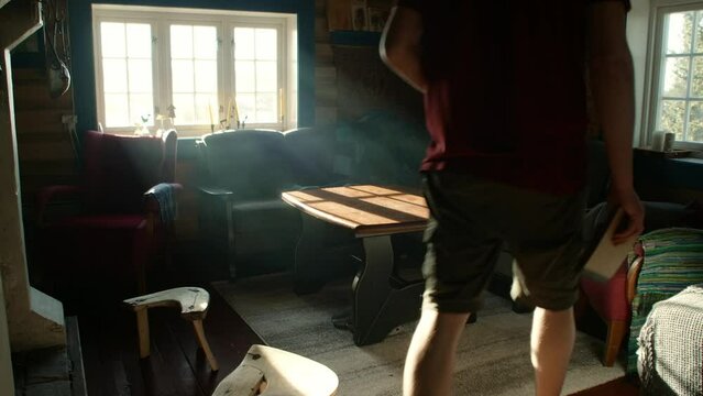 Young man entering living room in traditional norwegian cabin with a french press coffe brewing and a sketchbook in the morning harsh sun