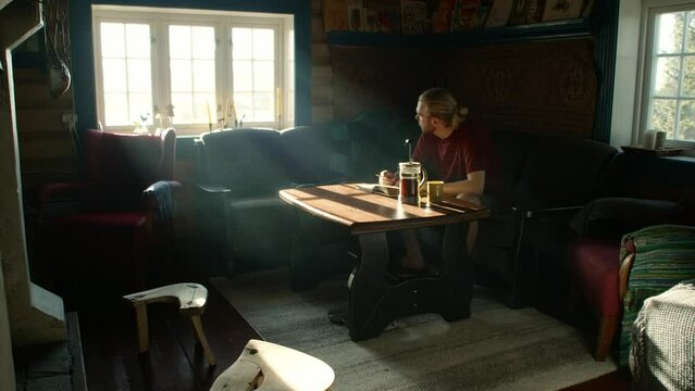Young man sitting in a traditional norwegian cabin thinking and looking out the window while brewing french press coffee
