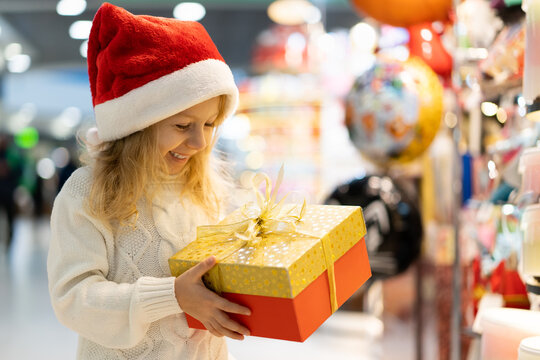 A Little Girl In A Christmas Hat With A New Year's Gift In Her Hands At A Showcase With Toys. Children Choose Gifts For Christmas. Black Friday Discounts