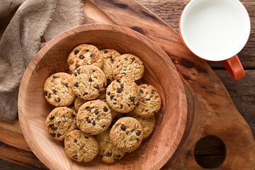 Chocolate chip cookies in wooden bowl with a cup of milk on the side, photographed overhead on wood...