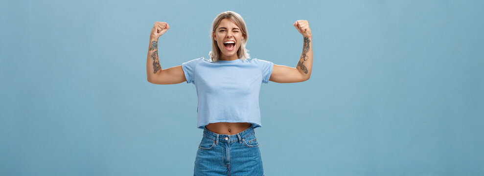 Strong women stand up and fight for rights. Portrait of happy entertained and cool young female athletic blonde with tattoos winking and smiling showing muscles and biceps over blue wall