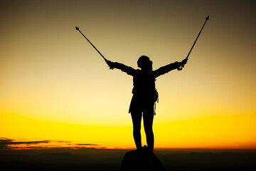 Silhouette of a hiker girl on a rock pedestal with hands up. Beautiful orange sunset.