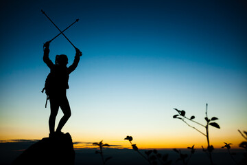 Silhouette of a hiker girl on a rock pedestal with hands up. Beautiful orange sunset on blue sky. Tree branches with leaves in the foreground.