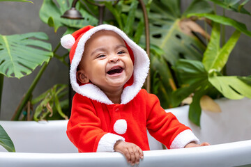 Portrait of African American little toddler baby girl in santa christmas dress smiling and playing inside the bathtub surrounding by green plant