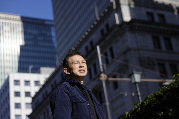 Middle-aged Asian man looking up at blue morning sky with high-rise business building in background.