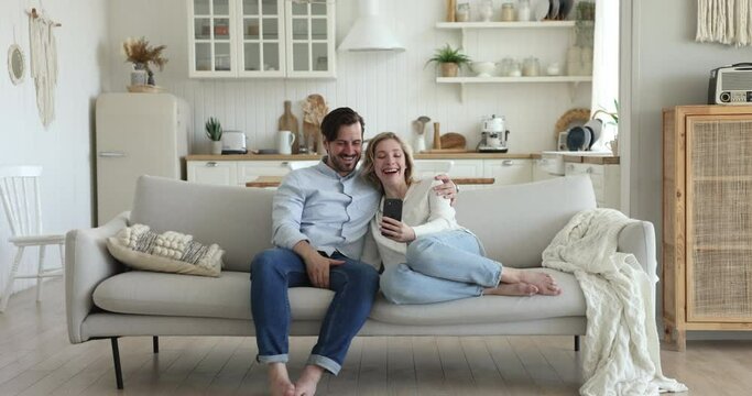 Cheerful millennial couple use new mobile application resting on cozy sofa with smartphone, staring at cellphone screen make self-portraits having fun on internet on leisure at home. Modern tech usage