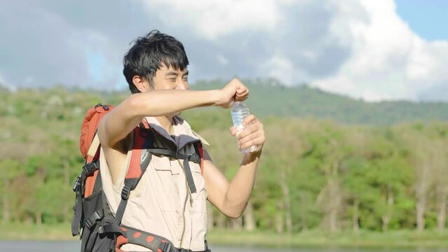 Young Asian man backpacker traveling in the forest. Male traveler holds a bottle of water. Healthy hiker drinking water in nature hike. Young man living active life, fun and smile.