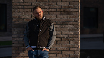 Caucasian bearded man in a bomber jacket leaned against a brick wall. 