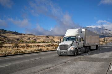 Bonnet industrial grade white big rig long hauler semi truck transporting cargo in dry van semi trailer running on the wide highway road with snow mountain and clouds on the background