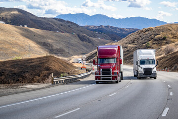 Burgundy and white two different big rig semi trucks with semi trailers climbing uphill on the winding mountain road going to mountain pass in California