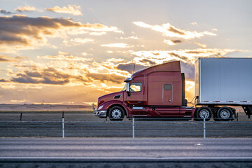 Burgundy big rig semi truck with grille guard and high cab spoiler transporting cargo in dry van semi trailer driving on the road at twilight