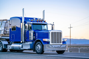 Gorgeous classic American blue big rig semi truck with chrome parts transporting cargo on flat bed semi trailer running on the evening flat road