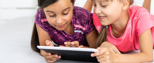 Two cute little girls are using a digital tablet and smiling while lying on bed in children's room