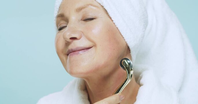 Senior woman in white bathrobe and towel on head doing spa skin care procedures using cosmetic roller for face massage on blue background