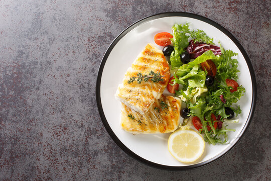 Spicy grilled cod fish fillet with fresh vegetable salad and leaf lettuce mix and lemon close-up in a plate on the table. Horizontal top view from above