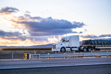Streamline big rig white semi truck with long hood transporting covered and fastened by slings cargo on flat bed semi trailer running on the road along the field at twilight sunset time