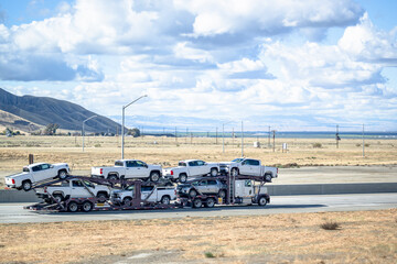 Fototapeta na wymiar Side view of the big rig car hauler semi truck transporting cars on the two level semi trailer driving on the highway road between fields