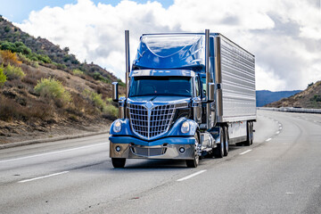 Stylish blue big rig semi truck with chrome parts transporting cargo in refrigerator semi trailer driving on the turning highway road in California mountain