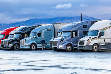 Truck driver walks towards his big rig semi truck in a row of other semi trucks with semi trailers standing on the truck stop parking lot