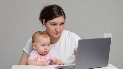 Brown-haired freelancer works on laptop holding baby daughter on lap against grey wall. Baby girl...