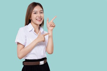 Portrait smiling of teen student girl of Asian ethnicity in university uniform pointing finger away on copy space isolated on light blue color background.
