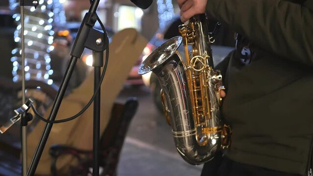 A street saxophonist in a city at night sparkling with Christmas lights and passing cars. The musician plays the saxophone earning money for food and improving the mood of people passing by
