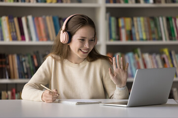 Happy cheerful gen Z high school student girl in headphones enjoying studying in library, consulting teacher on video call, attending online class, talking, waving hand hello at screen, smiling