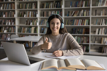 Happy confident student girl in headphones showing hand like sign, thumb up gesture, looking at camera, smiling, working in university library, writing essay, article. Head shot portrait