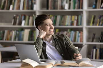 Happy dreamy student guy working on scientific article, study research in library, writing notes at...