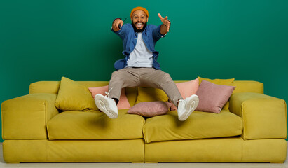 Happy young man using remote control while jumping on the couch against green background