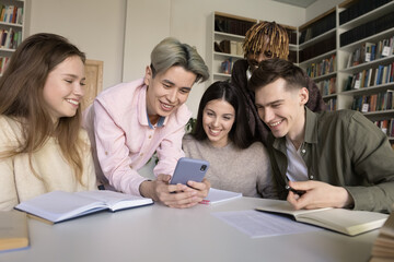 Diverse team of cheerful teenager students using smartphone together, making video call in university library, watching learning video, online lesson, consulting teacher, smiling