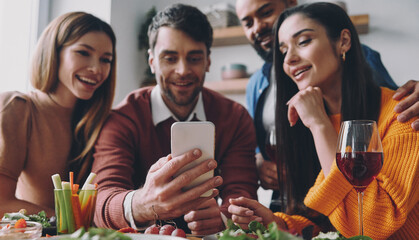 Group of happy young people looking at smart phone while enjoying dinner at home together