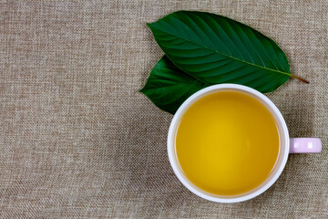 Mitragyna Speciosa Korth or kratom tea in white cup wtih green leaf on sackcloth, top view, copy space.