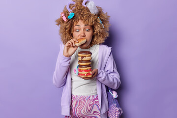 Surprised curly haired pregnant woman eats bun stares at delicious sweet doughnuts being hungry all time carries net bag on shoulder dressed casually isolated over purple background. Unhealthy eating