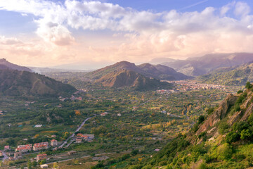 Fototapeta premium beautiful landscape with amazing view from highland with green slopes and bushes to a valley with town, road and nice mountains and scenic cloudy sunset on background
