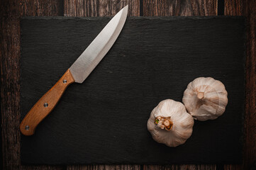 Garlic on a black slate cutting board with a knife with a wooden handle. On a dark wooden table.