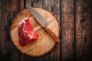 A piece of red beef on a rustic wooden table with a sharp knife. Top down view.