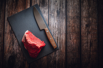 A piece of raw beef on a slate cutting board with a knife. Wooden table, top down view, flat lay.